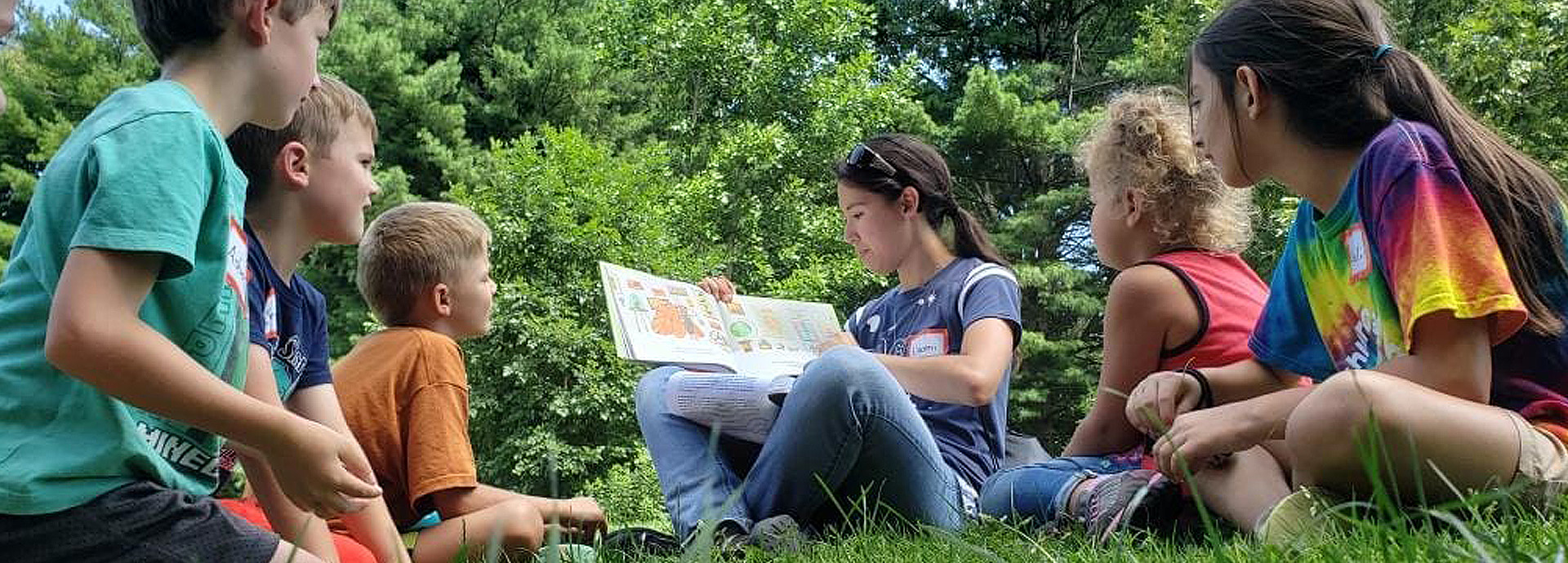 Slideshow Image - A staff member sitting on the grass with a group of small children and reading a book about butterflies to them